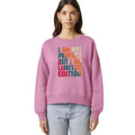 A woman modeling a pink Stanley/Stella Alma crewneck sweatshirt with a colorful graphic text print that reads 'I AM NOT PERFECT BUT I AM LIMITED EDITION' in teal, orange, and yellow tones, paired with light blue jeans against a plain white background.