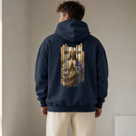 A man viewed from behind wearing a navy blue hoodie with a large graphic print of Jesus Christ on the back, paired with cream-colored pants, in a neutral indoor setting.