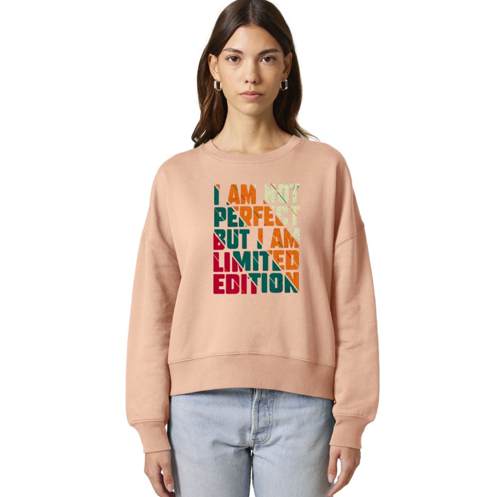 A woman modeling a peach-colored crewneck sweatshirt with a colorful graphic text print that reads 'I AM NOT PERFECT BUT I AM LIMITED EDITION' in vertical stacked letters, paired with light blue jeans against a plain white background.