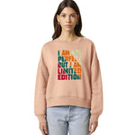 A woman modeling a peach-colored crewneck sweatshirt with a colorful graphic text print that reads 'I AM NOT PERFECT BUT I AM LIMITED EDITION' in vertical stacked letters, paired with light blue jeans against a plain white background.
