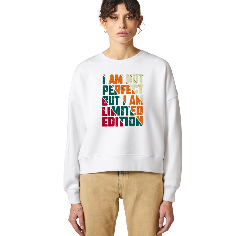 A model wearing a white crewneck sweatshirt with a colorful text graphic that reads 'I AM NOT PERFECT BUT I AM LIMITED EDITION' in a retro block-letter design. The model is also wearing beige pants, set against a plain white studio background.
