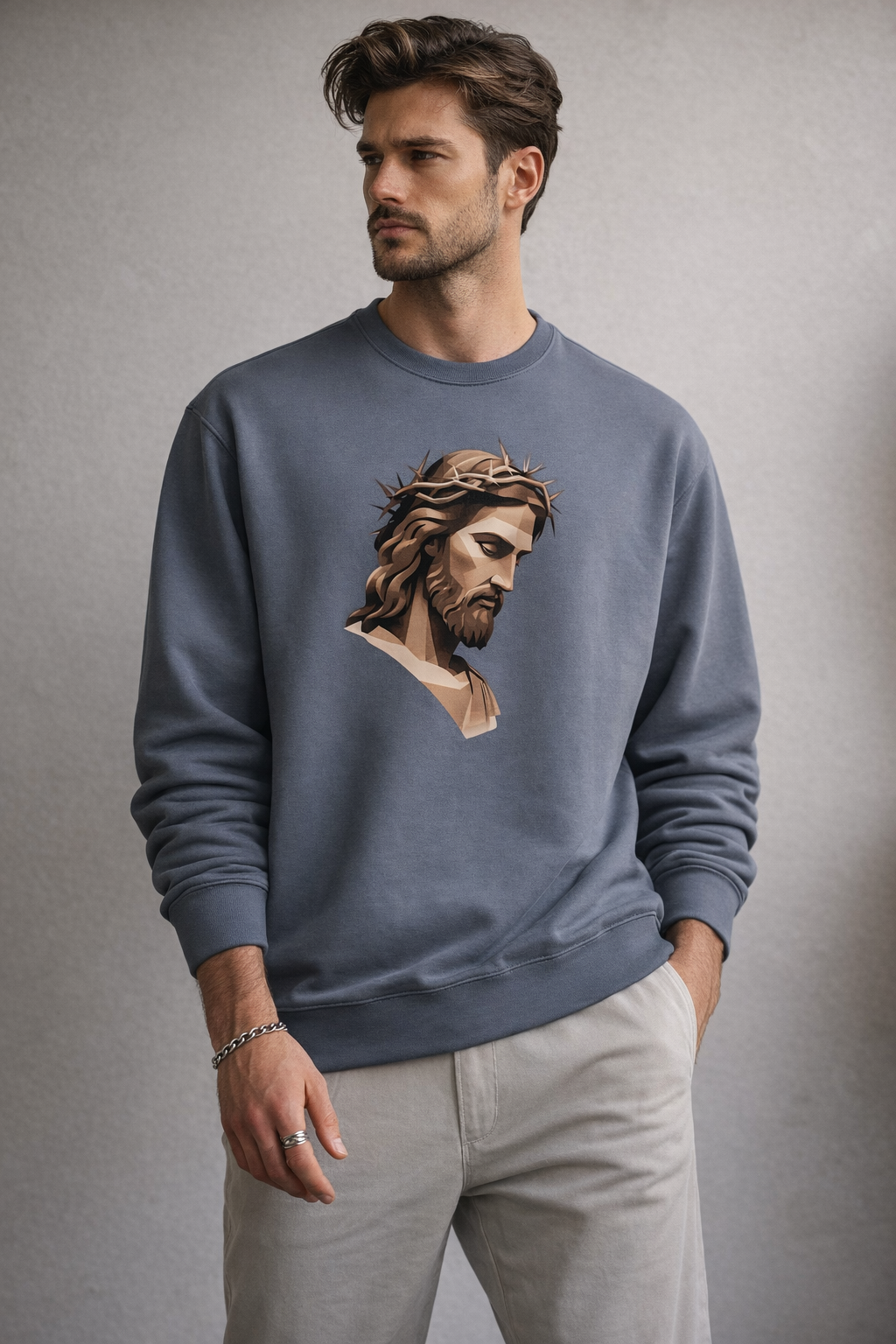 A man with tousled hair and a beard wears a blue sweatshirt featuring an illustrated image of Jesus with a crown of thorns. He stands against a plain, light gray background with one hand in his pocket.