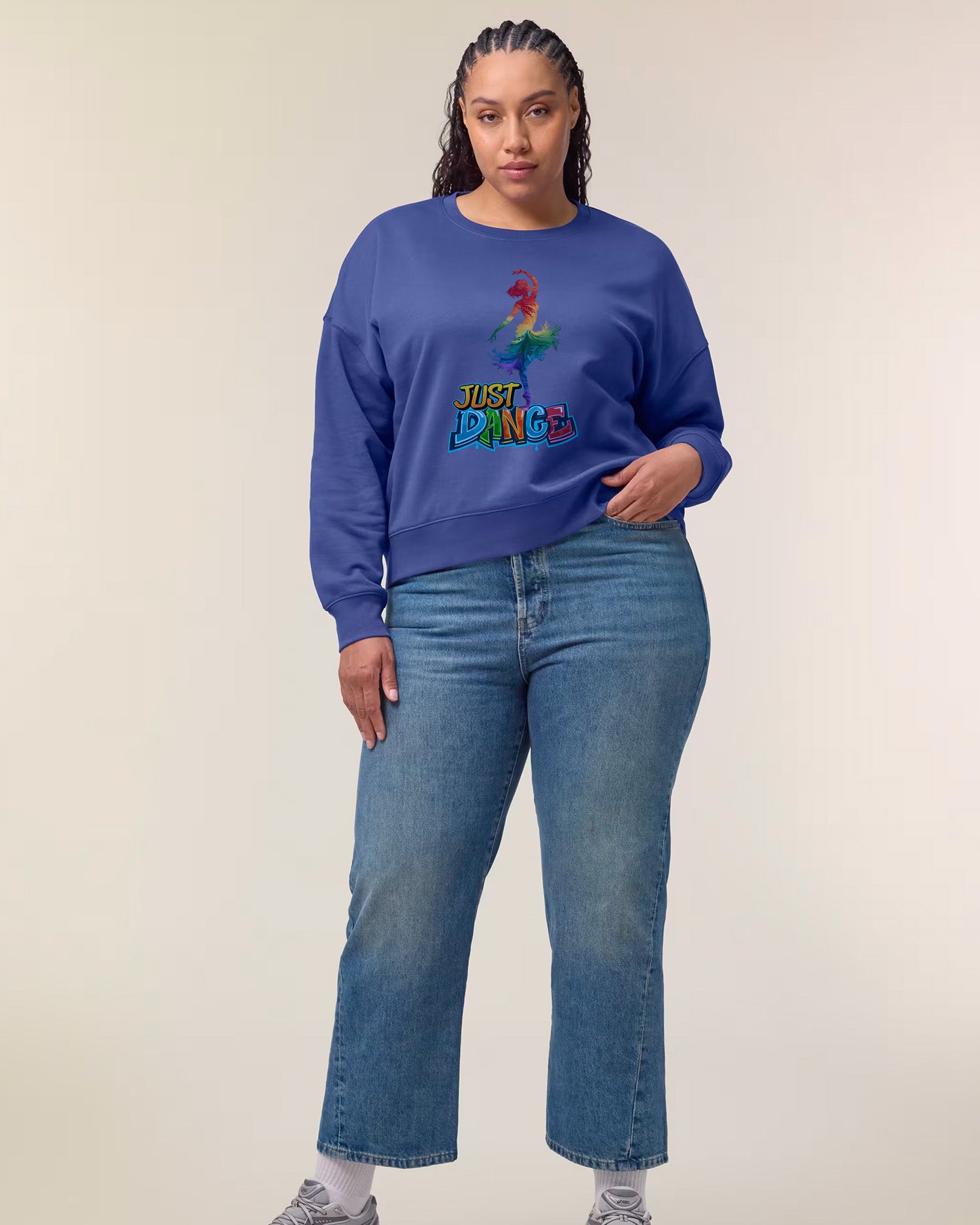 A woman with braided hair wears a blue sweatshirt featuring a colorful dinosaur and the text "Just Dinge." She is also wearing blue jeans and white sneakers, standing against a plain light background.