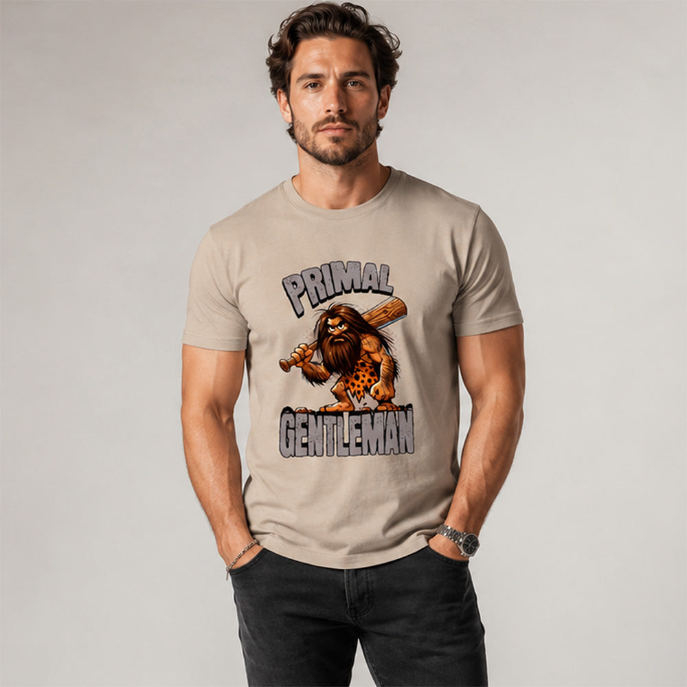 A man wearing a beige graphic t-shirt with a 'Primal Gentleman' caveman design, paired with black jeans, standing against a neutral gray studio background.