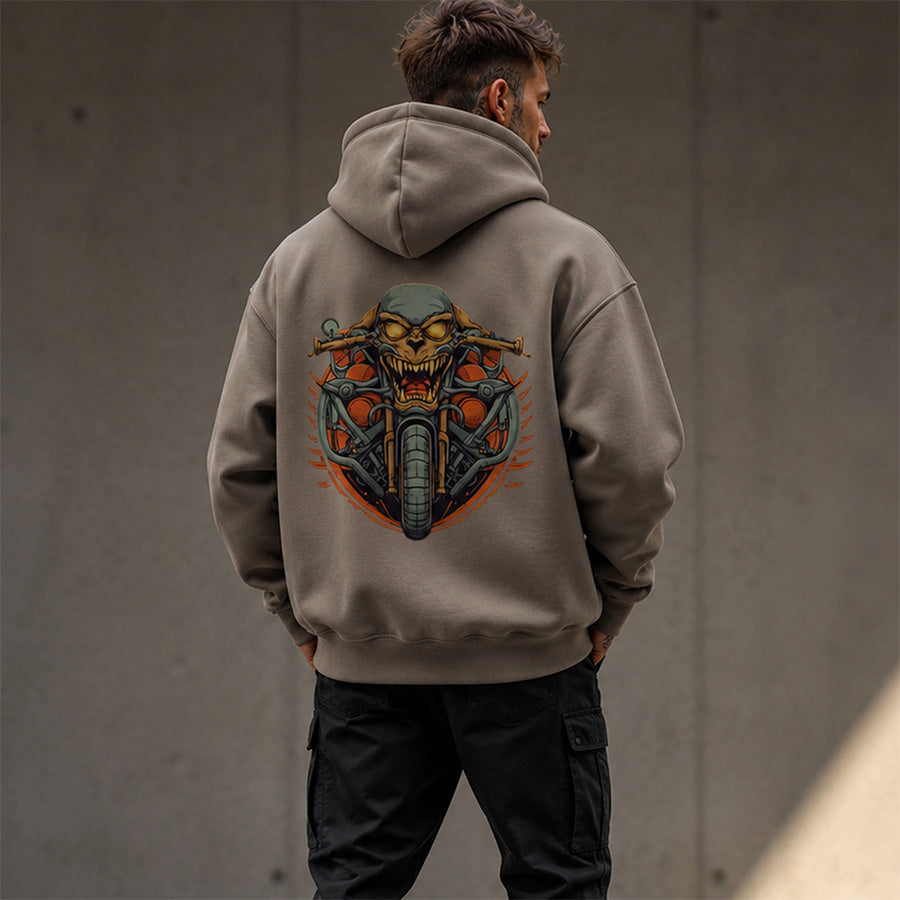 A man viewed from behind wearing a taupe hoodie with a large graphic print of a skull on a motorcycle on the back, paired with black cargo pants against a concrete wall.