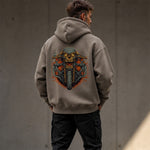 A man viewed from behind wearing a taupe hoodie with a large graphic print of a skull on a motorcycle on the back, paired with black cargo pants against a concrete wall.
