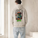 A man viewed from behind wearing a light beige hoodie with a large colorful graphic on the back featuring the words 'FLYING RICH' and a seated green alien smoking a mushroom, paired with light blue jeans in a minimalist indoor setting.