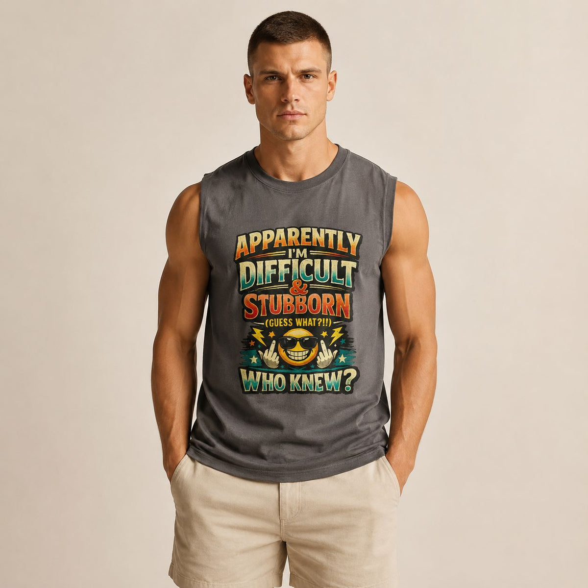 A muscular man wearing a dark gray sleeveless muscle tank top with a colorful graphic text print that reads 'APPARENTLY IN DIFFICULT & STUBORN (GUESS WHAT?!)' and 'WHO KNEW?'. He is also wearing beige shorts, standing against a solid light beige background.