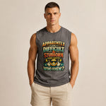 A muscular man wearing a dark gray sleeveless muscle tank top with a colorful graphic text print that reads 'APPARENTLY IN DIFFICULT & STUBORN (GUESS WHAT?!)' and 'WHO KNEW?'. He is also wearing beige shorts, standing against a solid light beige background.