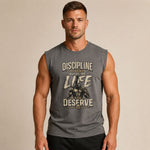 A muscular man wearing a heather gray sleeveless muscle tank top with a graphic print that reads 'DISCIPLINE TODAY BUILDS THE LIFE YOU DESERVE' and shows a bodybuilder illustration. The background is a solid warm beige studio backdrop.