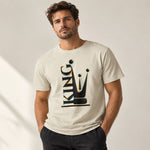 A man wearing a light beige crewneck t-shirt with a stylized graphic of a crown and the word 'KING' in black and gold. He is standing against a neutral light beige wall with soft shadows.