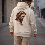 A man wearing a cream-colored hoodie with an artistic image of Jesus wearing a crown of thorns printed on the back, standing in an empty city street.