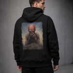 A man wearing a black hoodie stands with his back to the camera. The hoodie features artwork of a red-skinned, eerie humanoid figure covering its mouth amid a dark, surreal landscape.