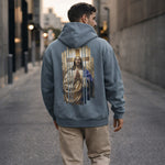A man stands in an urban alley wearing a blue hoodie with an image of Jesus praying in front of a cross printed on the back.