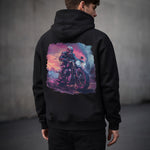 Man wearing a black hoodie with a colorful graphic of a person on a motorcycle on the back against a gray background.