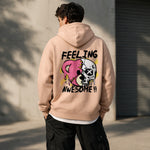 A man wearing a beige hoodie with colorful graffiti art and the words "FEELING AWESOME!!" on the back stands outdoors by a concrete wall and a metal garage door.