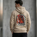A man wearing a beige hoodie stands with his back to the camera. The hoodie features a large, colorful graphic of a geisha with fans and flowers on the back. The background is blurred and gray.