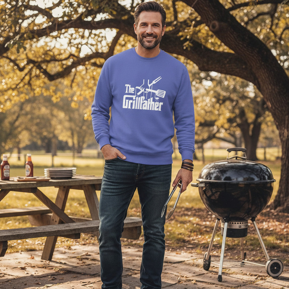 Man wearing a blue sweatshirt with 'The Grillfather' text, standing next to a barbecue grill outdoors.