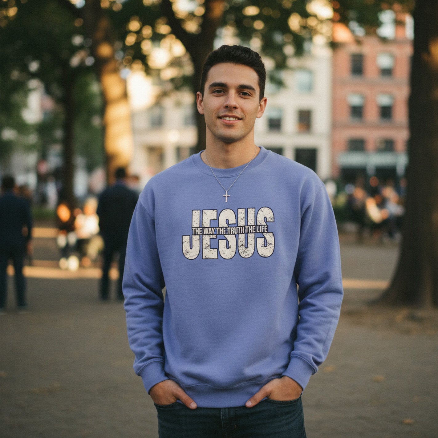 A young man stands outdoors in a park, wearing a light blue sweatshirt that reads "JESUS THE WAY THE TRUTH THE LIFE" in large letters. He also wears a silver cross necklace and smiles at the camera, with trees and buildings in the background.