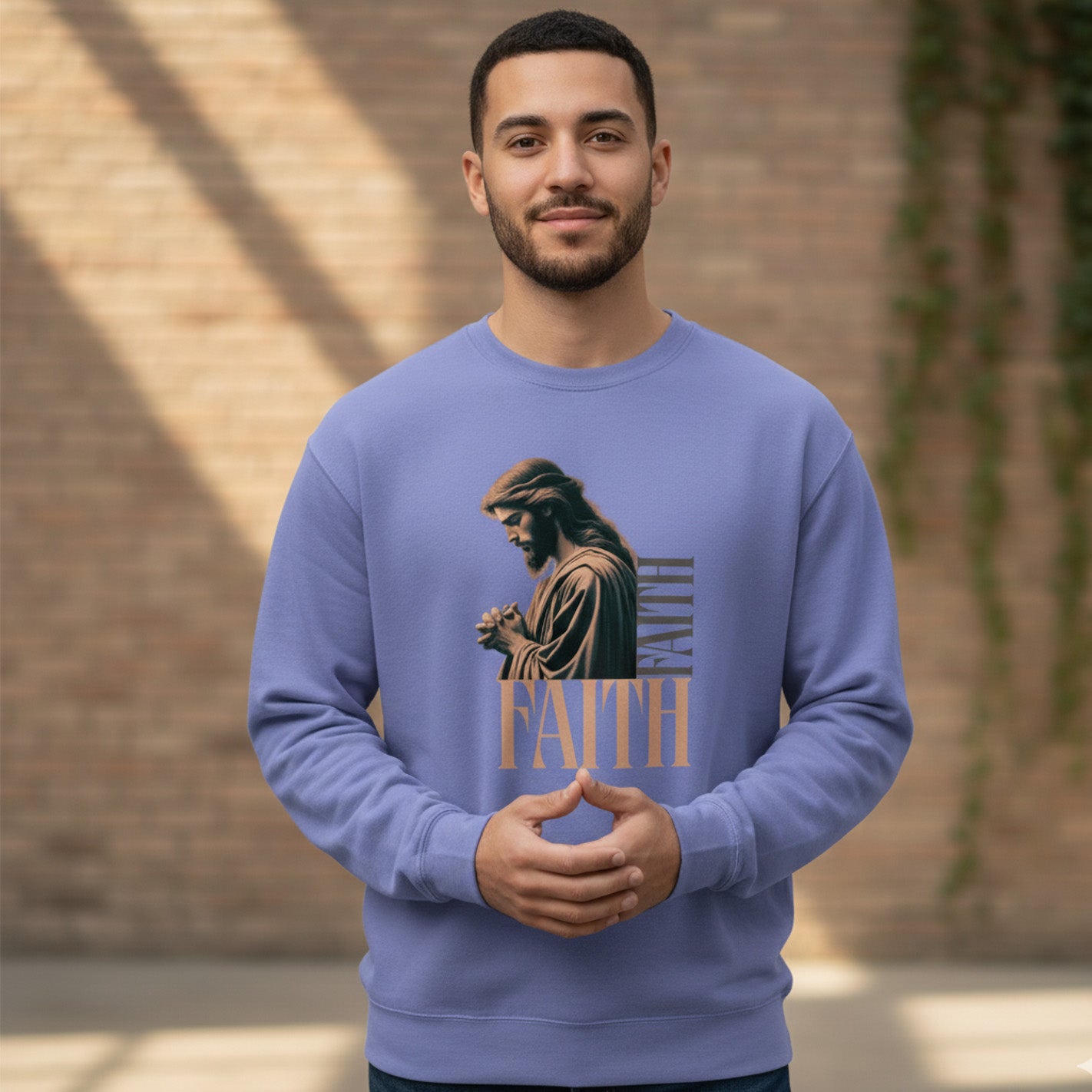 A man stands indoors wearing a light purple sweatshirt with an image of Jesus praying and the word "FAITH" printed below. The background features a brick wall and some green plants.