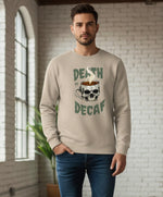 A man stands indoors wearing a light beige sweatshirt with a graphic of a coffee cup shaped like a skull and the words “Death in Decaf.” He has short hair and a beard, and the background features a white brick wall and a window.