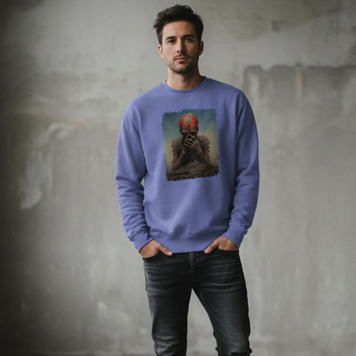 A man stands indoors wearing a purple sweatshirt with an artistic print of a red-faced figure holding its head, paired with dark jeans, against a textured gray wall.