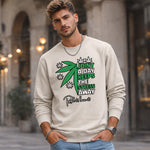 A young man stands outdoors wearing a beige sweatshirt with a graphic of a cannabis leaf and text that reads, "Joint a day keeps the stress away. Roll it, love it." He has short hair, a beard, and wears a chain necklace.