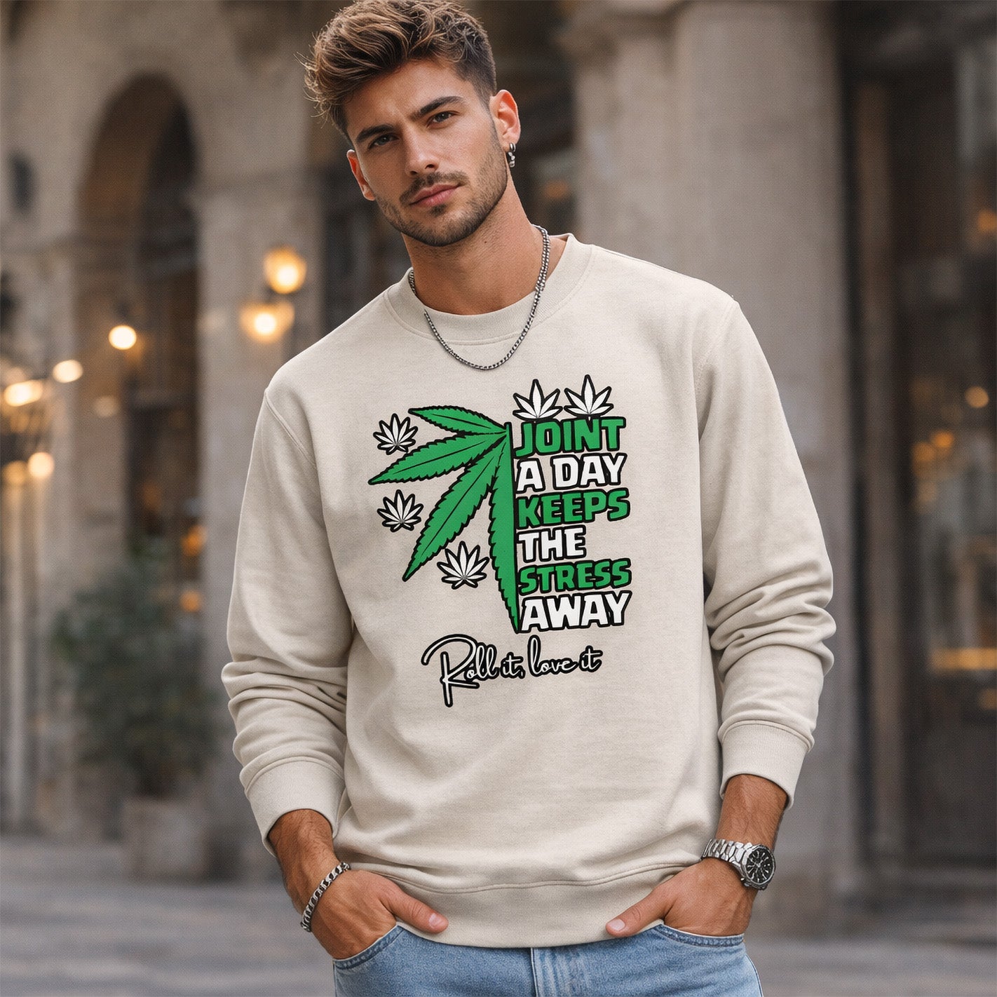 A young man stands outdoors wearing a beige sweatshirt with a graphic of a cannabis leaf and text that reads, "Joint a day keeps the stress away. Roll it, love it." He has short hair, a beard, and wears a chain necklace.