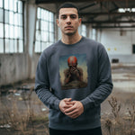 A man stands in an abandoned industrial building, wearing a gray sweatshirt with artwork depicting a distressed figure covering its face. Dry plants and broken windows are visible in the background.