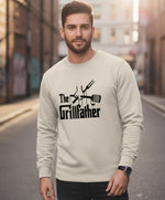 A man with a beard wearing a light-colored sweatshirt that says "The Grillfather" with images of grilling utensils stands in an urban alleyway, looking at the camera.