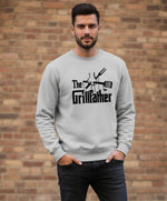 A man stands against a brick wall, wearing a light gray sweatshirt that says "The Grillfather" with grilling utensils graphic. He has short dark hair, a trimmed beard, and his hands in his pockets.