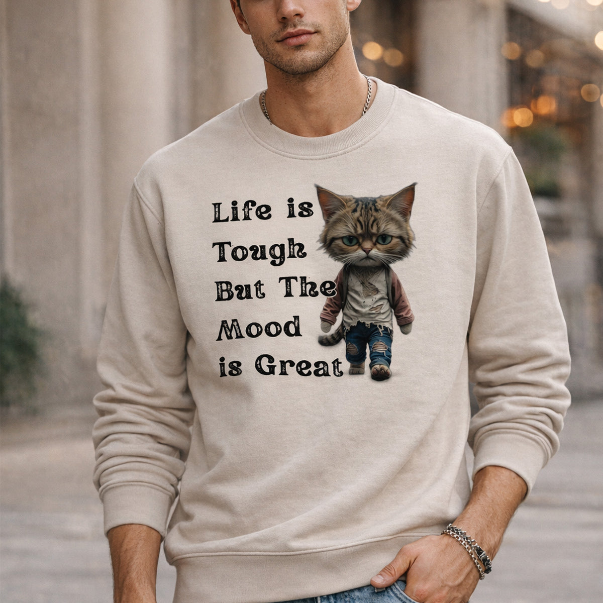 A man wearing a beige sweatshirt that features a cartoon cat dressed in jeans and a sweater, with the text “Life is Tough But The Mood is Great” printed beside the cat.