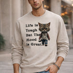 A man wearing a beige sweatshirt that features a cartoon cat dressed in jeans and a sweater, with the text “Life is Tough But The Mood is Great” printed beside the cat.