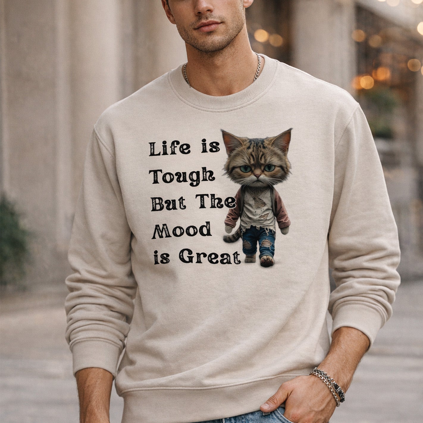 A man wearing a beige sweatshirt that features a cartoon cat dressed in jeans and a sweater, with the text “Life is Tough But The Mood is Great” printed beside the cat.