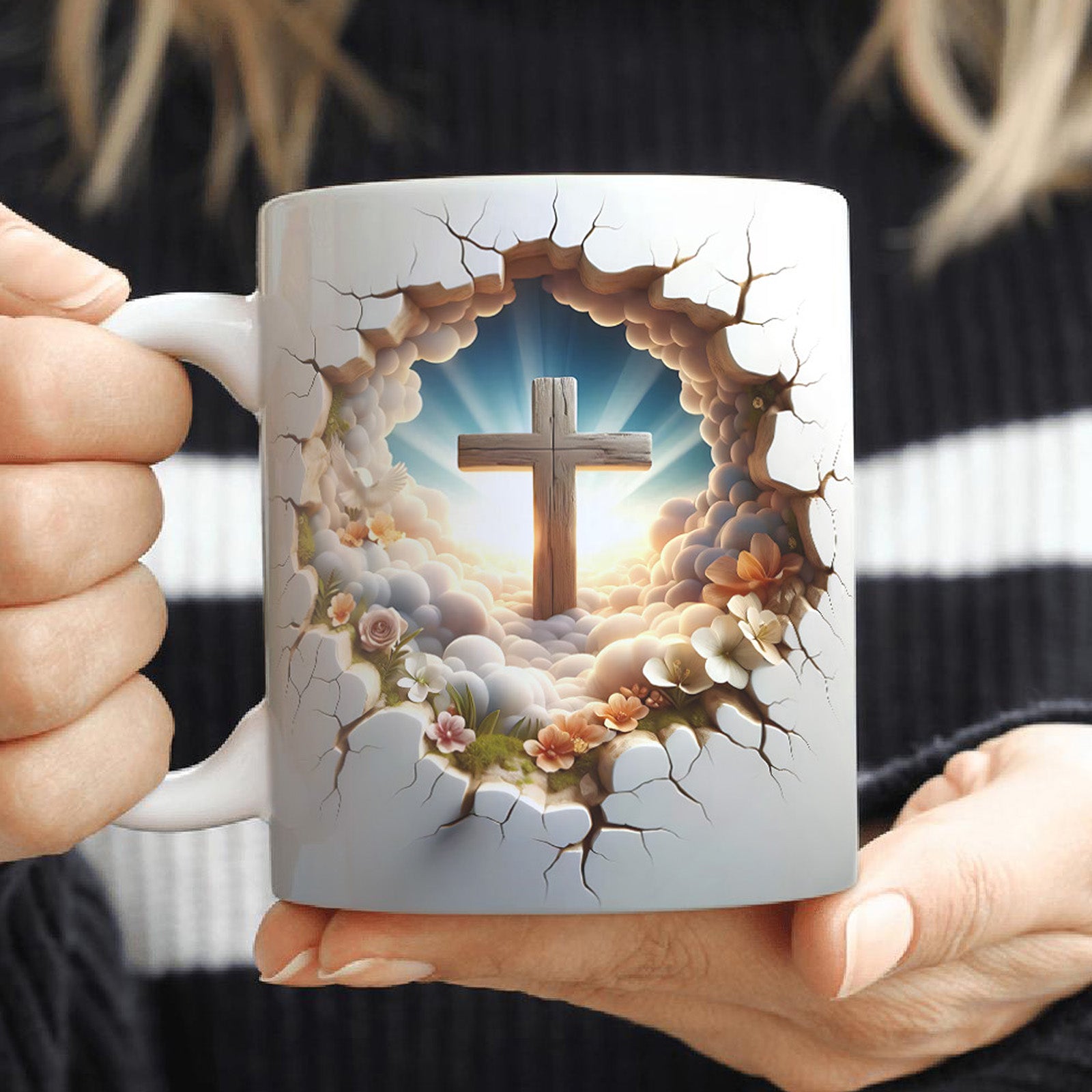 A woman holds a white mug featuring an image of a wooden cross surrounded by clouds, sunlight, and flowers, with a cracked design creating a 3D effect on the surface.