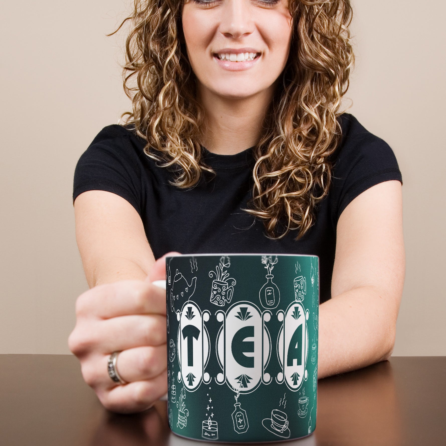 A woman with curly hair and a black shirt smiles while holding a dark green mug with the word "TEA" and whimsical white illustrations on it.