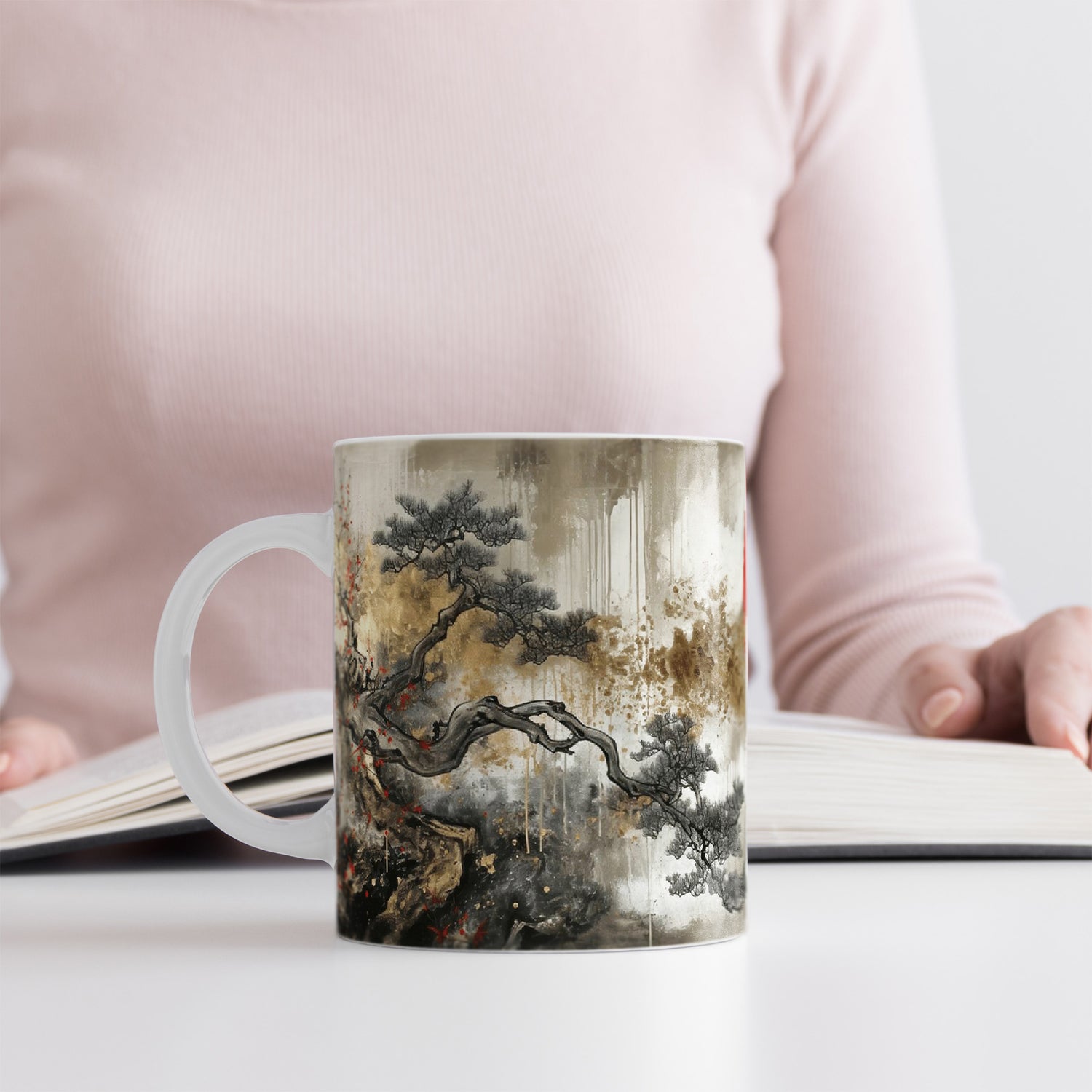 Woman in a light pink top reads a book at a white table. In front of them is a mug decorated with traditional Asian-style art featuring a tree with twisted branches and muted earthy colors.