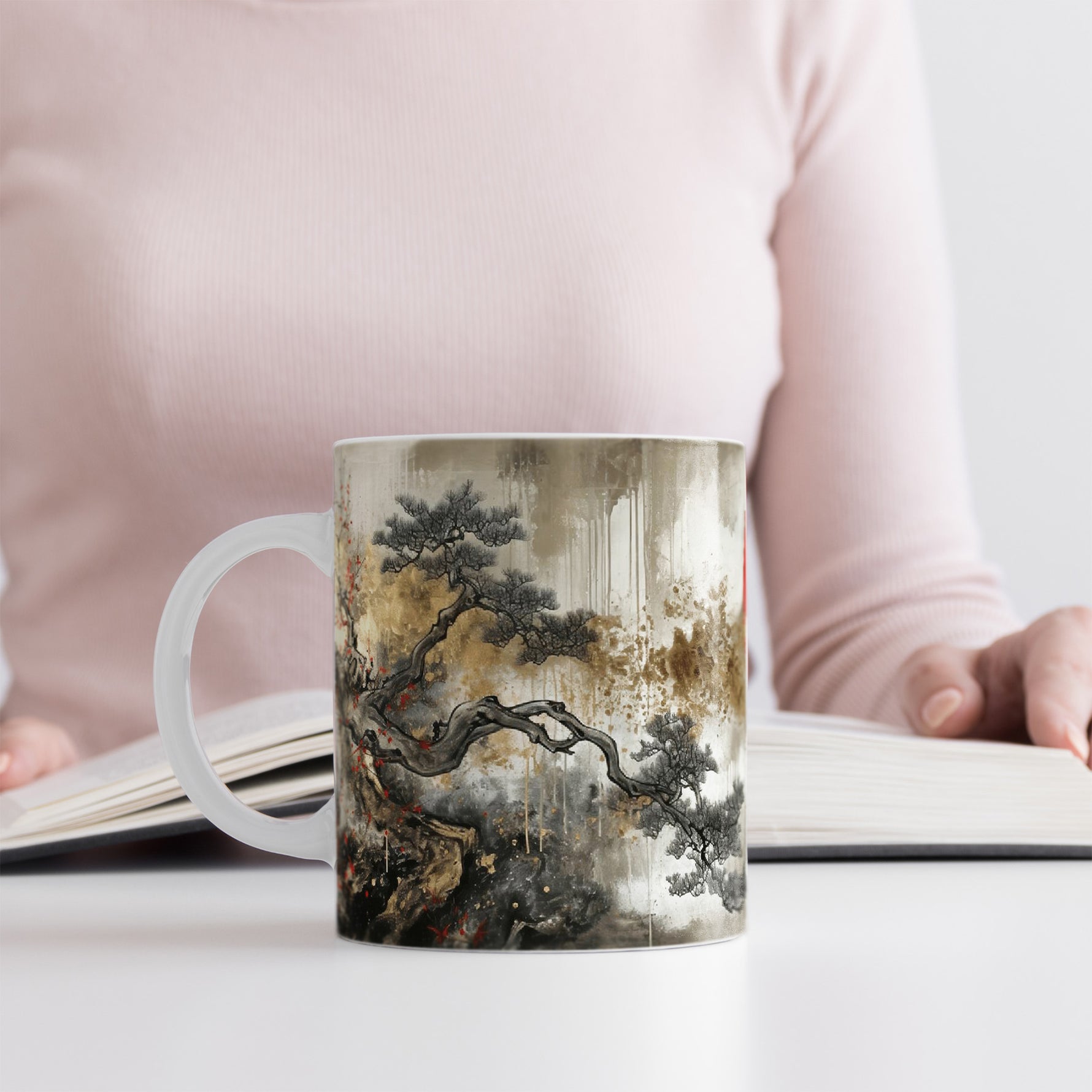 Woman in a light pink top reads a book at a white table. In front of them is a mug decorated with traditional Asian-style art featuring a tree with twisted branches and muted earthy colors.
