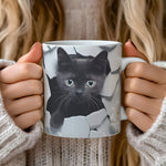 A woman in a cozy sweater holds a mug featuring an illustration of a black kitten peeking through a cracked wall.