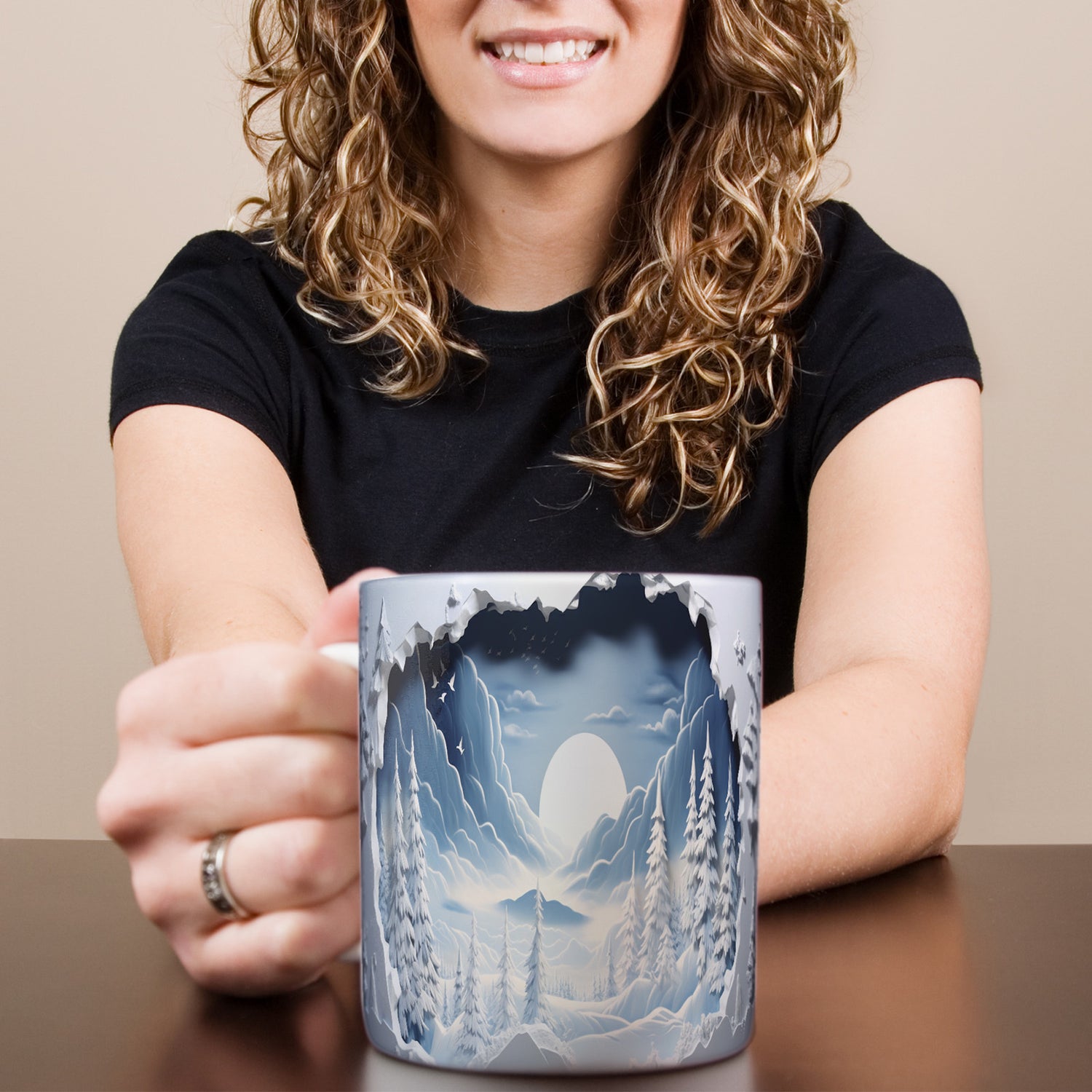 A smiling woman with curly hair holds a mug featuring a winter landscape with snow-covered trees, mountains, and a full moon against a dark sky.