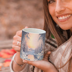 A smiling woman in a cozy scarf holds a mug decorated with a snowy winter landscape, featuring pine trees and a river at sunset. Autumn leaves are scattered on the wooden surface in the background.