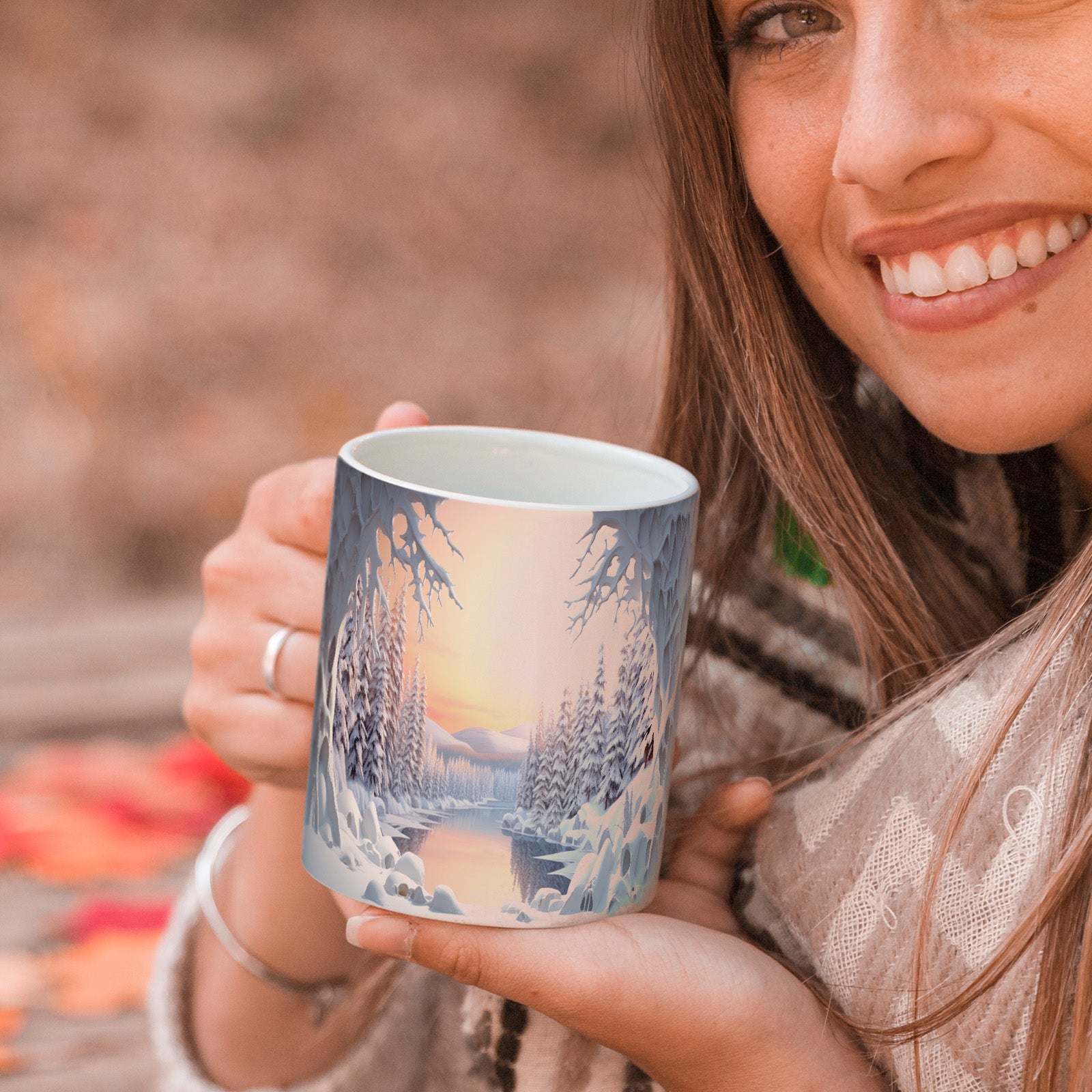 A smiling woman in a cozy scarf holds a mug decorated with a snowy winter landscape, featuring pine trees and a river at sunset. Autumn leaves are scattered on the wooden surface in the background.