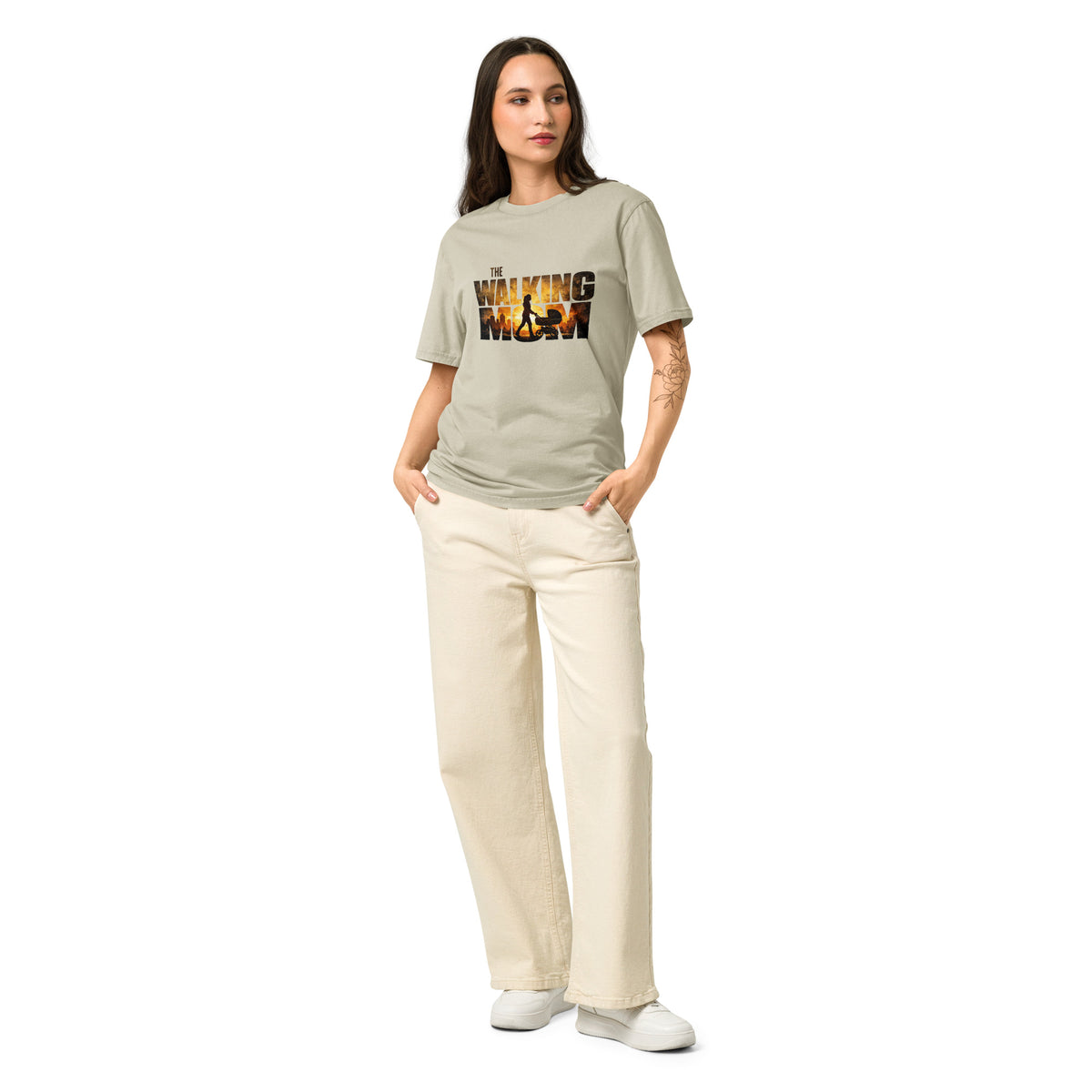 A woman modeling a light sage green graphic t-shirt with 'WAVING WOMAN' printed in a fiery orange and yellow pattern, paired with cream-colored pants and white sneakers, set against a plain white background.