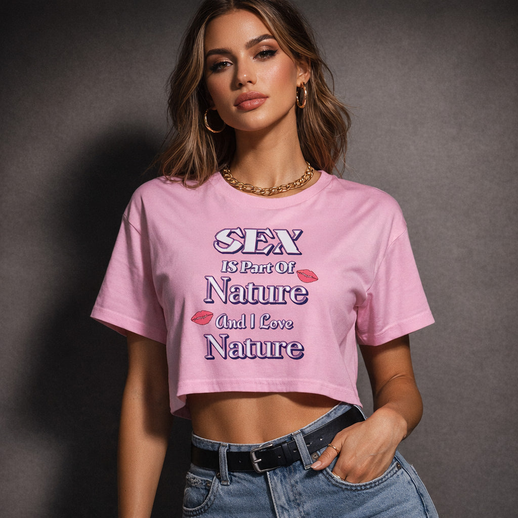A woman wearing a pink crop top with the text "SEX is part of Nature and I love Nature" stands confidently with one hand on her hip, sporting high-waisted jeans, gold hoop earrings, and a chain necklace.