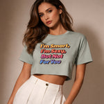 A woman with long brown hair wearing a light gray crop top with colorful text that reads, "I'm Smart, I'm Sexy, But Not For You," paired with high-waisted white pants, stands against a neutral background.