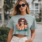 A woman wearing sunglasses and a crop top with a graphic of a retro-style woman, the word "Respectfully," and white high-waisted pants, stands outdoors with palm trees and buildings in the background.