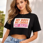 A woman wearing hoop earrings and a black cropped t-shirt with the words "MY BODY MY CHOICE MY RULES" in bold orange, pink, and white text stands outdoors, posing confidently with one hand on her hip.