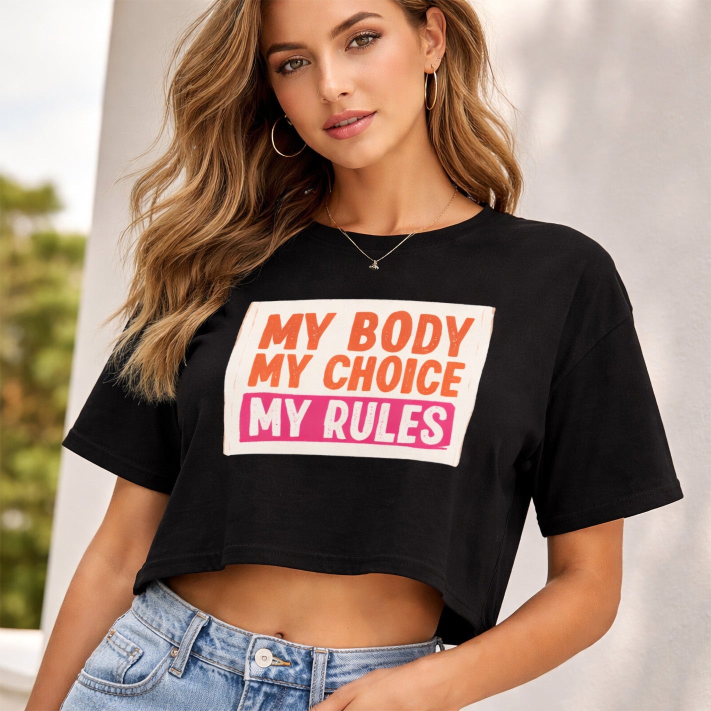 A woman wearing hoop earrings and a black cropped t-shirt with the words "MY BODY MY CHOICE MY RULES" in bold orange, pink, and white text stands outdoors, posing confidently with one hand on her hip.