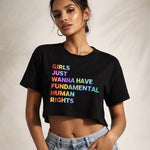 A woman wears a black crop top with the words “Girls just wanna have fundamental human rights” printed in colorful, bold letters. She stands confidently against a neutral background.