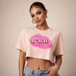 A woman wearing hoop earrings and a light pink crop top with a large pink lip print and the word "b*tch!" printed on it, paired with blue jeans, standing against a neutral background.